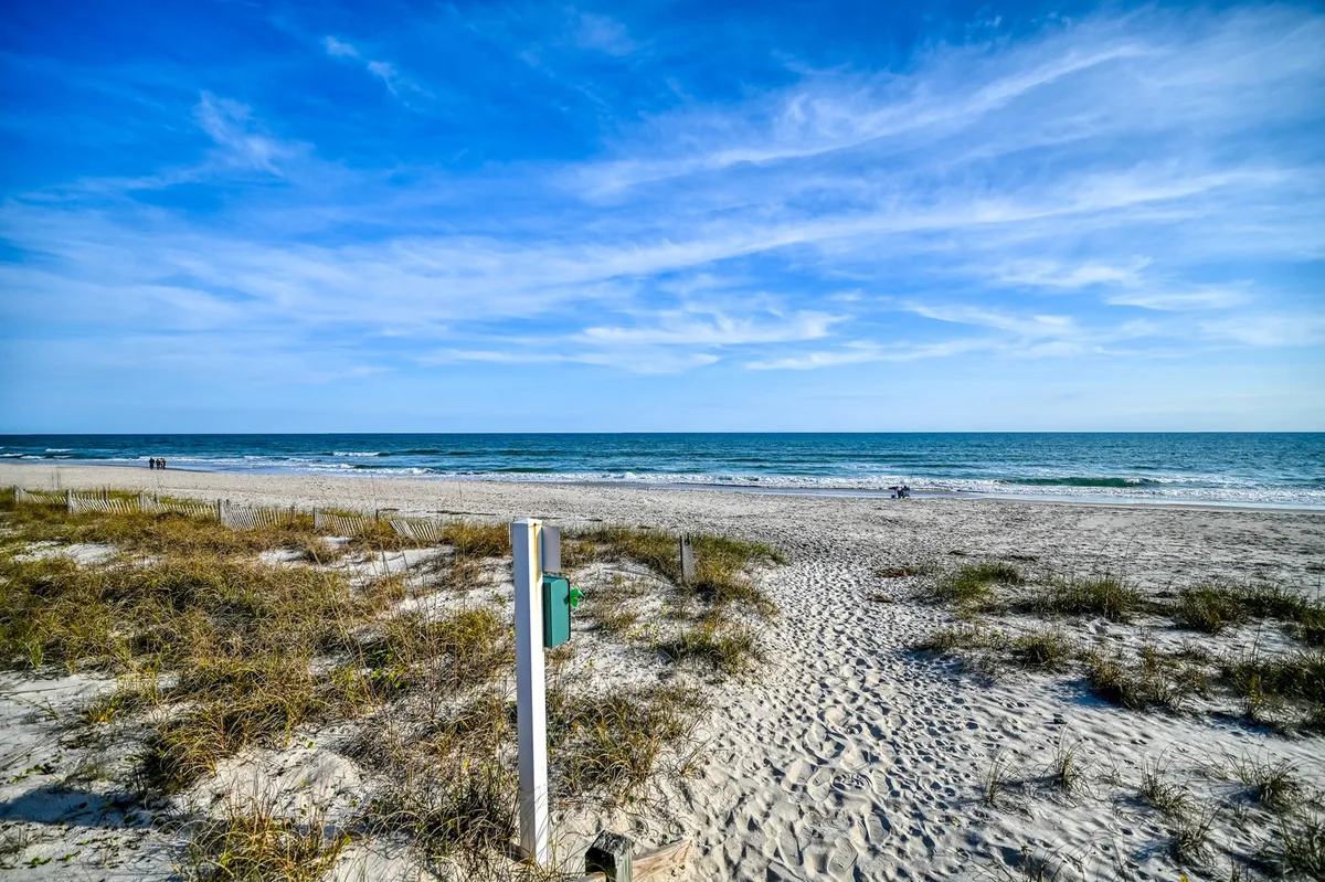 A natural beach path leads through dune grasses to a serene ocean vista under a cloudy sky. The scene evokes peaceful beachcombing, sunset strolls, and the natural beauty of the coastline.