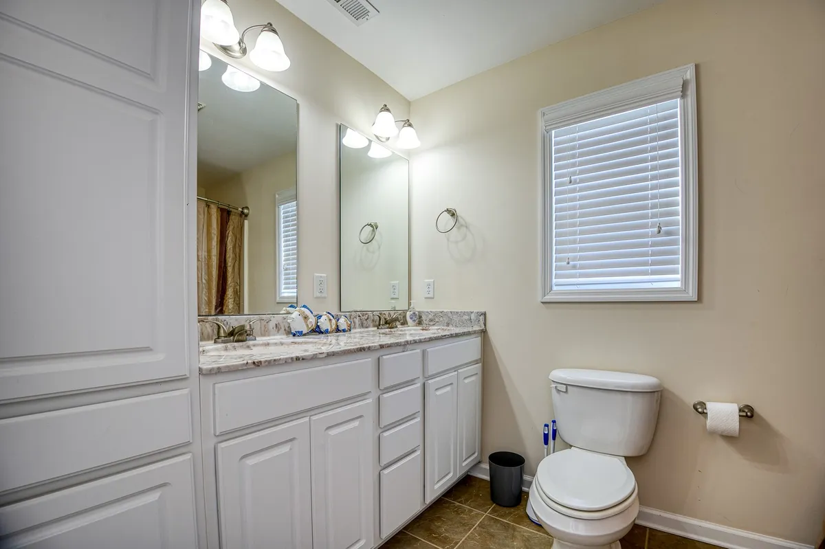A serene bathroom setting with a double sink vanity, large mirrors, modern light fixtures, and a window with blinds. The space is orderly and calming, ideal for self-care and relaxation.