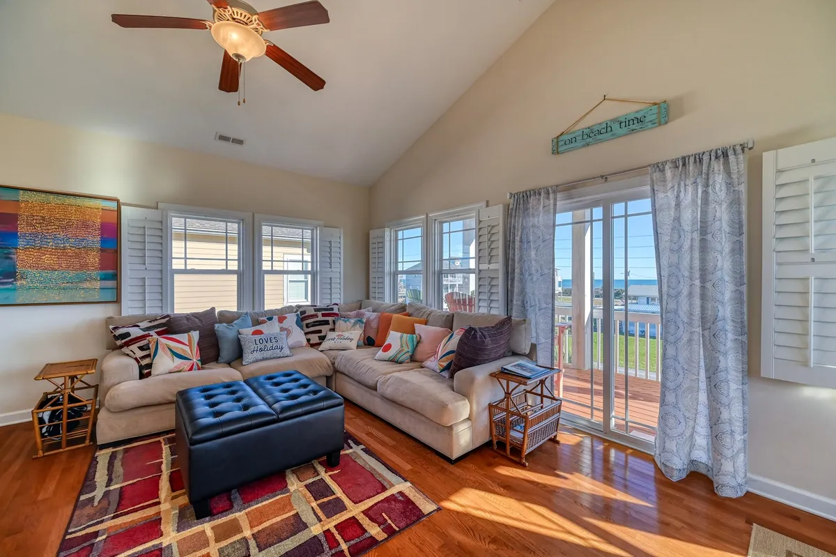 A large, inviting sectional sofa decorated with colorful throw pillows anchors the spacious open-concept living area. Large windows flood the space with natural light, offering views of the outdoors. The room is accented by a patterned rug and ceiling fan, creating a cozy atmosphere perfect for family gatherings, movie nights, or relaxing after a day at the beach.
