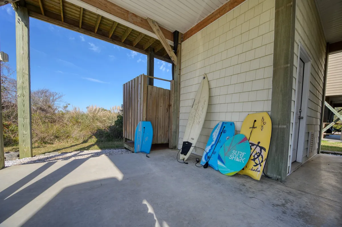 This exterior image features an outdoor shower beside a patio area with surfboards resting nearby. The setup is casual and functional, perfect for rinsing off after beach adventures with family, friends, or furry companions.