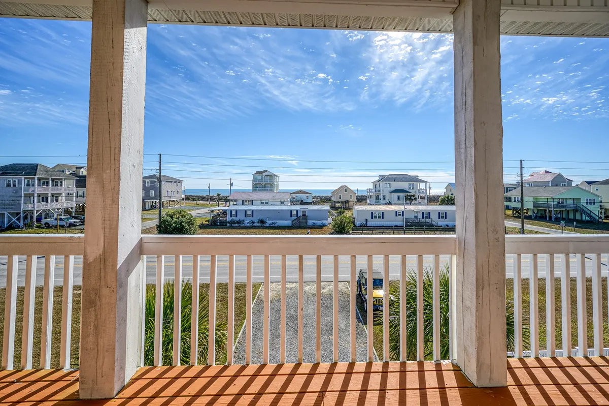 A relaxed deck setup overlooks the ocean, with deck railing, comfortable seating, and surrounding beach houses in the frame. Clear blue skies and the expansive ocean view create a laid-back, inviting atmosphere.