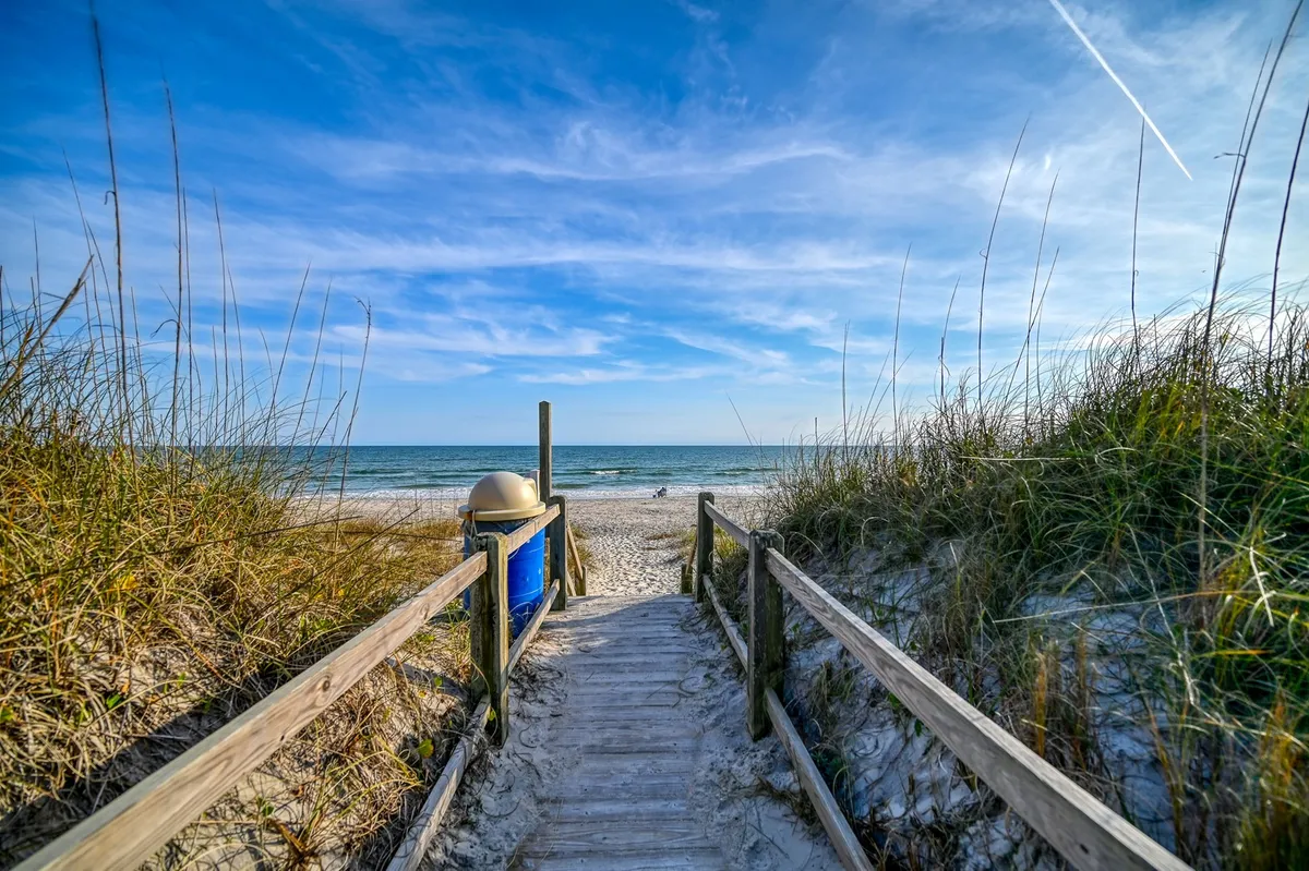 A charming wooden walkway leads from a classic blue beach box home through dune grasses to the expansive shoreline. The ocean is visible in the background, with soft morning light and gentle waves beckoning beachgoers. The scene captures the easy, direct access from the house to the sandy beach, inviting families for sunbathing, swimming, and sandcastle building.