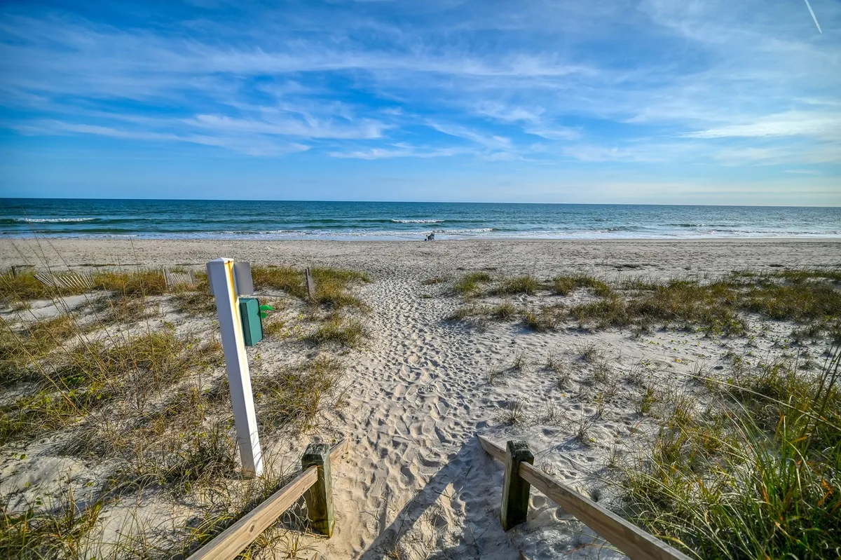 A sandy pathway leads to the beach, with distant figures on the shore and the ocean waves beyond. Children are seen building sandcastles, and the scene exudes a relaxed, family-friendly, and playful mood, perfect for sunbathing and beach adventures.