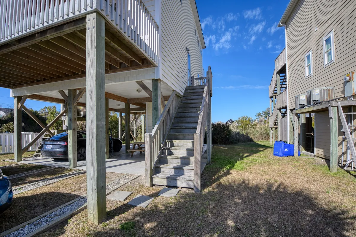 This exterior image showcases the open parking area beneath the house, a staircase leading to the upper deck, and adjacent houses visible in the background. The open space under the house is well-lit and clearly structured, making it easy to imagine parking multiple vehicles and accessing the main entrance after a day at the beach.