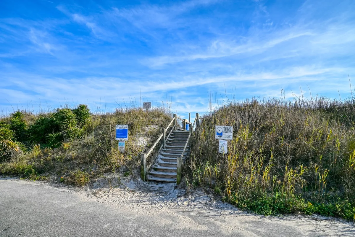 A wooden staircase leads over dune grass toward a pristine beach, with clear blue sky above. Signs indicate beach access, and the area is flanked by natural vegetation, evoking a sense of easy exploration and connection to nature.