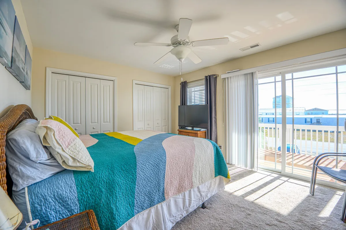 A bright and inviting bedroom with a colorful quilt on the bed, large windows that flood the space with natural light, a wicker bed frame, and a TV unit. The setup exudes comfort and modern amenities, making it ideal for relaxation or enjoying a quiet evening.