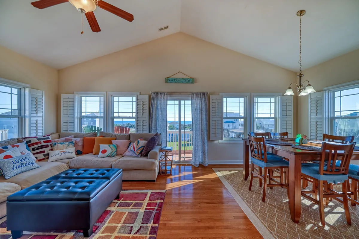 A spacious, light-filled living and dining area featuring a large, cozy sectional sofa adorned with colorful pillows, a welcoming dining table with ample seating, and expansive windows offering stunning sunset water views. The room's inviting atmosphere is enhanced by a stylish rug and the soft glow from natural light, perfect for gathering, relaxing, or celebrating with loved ones.