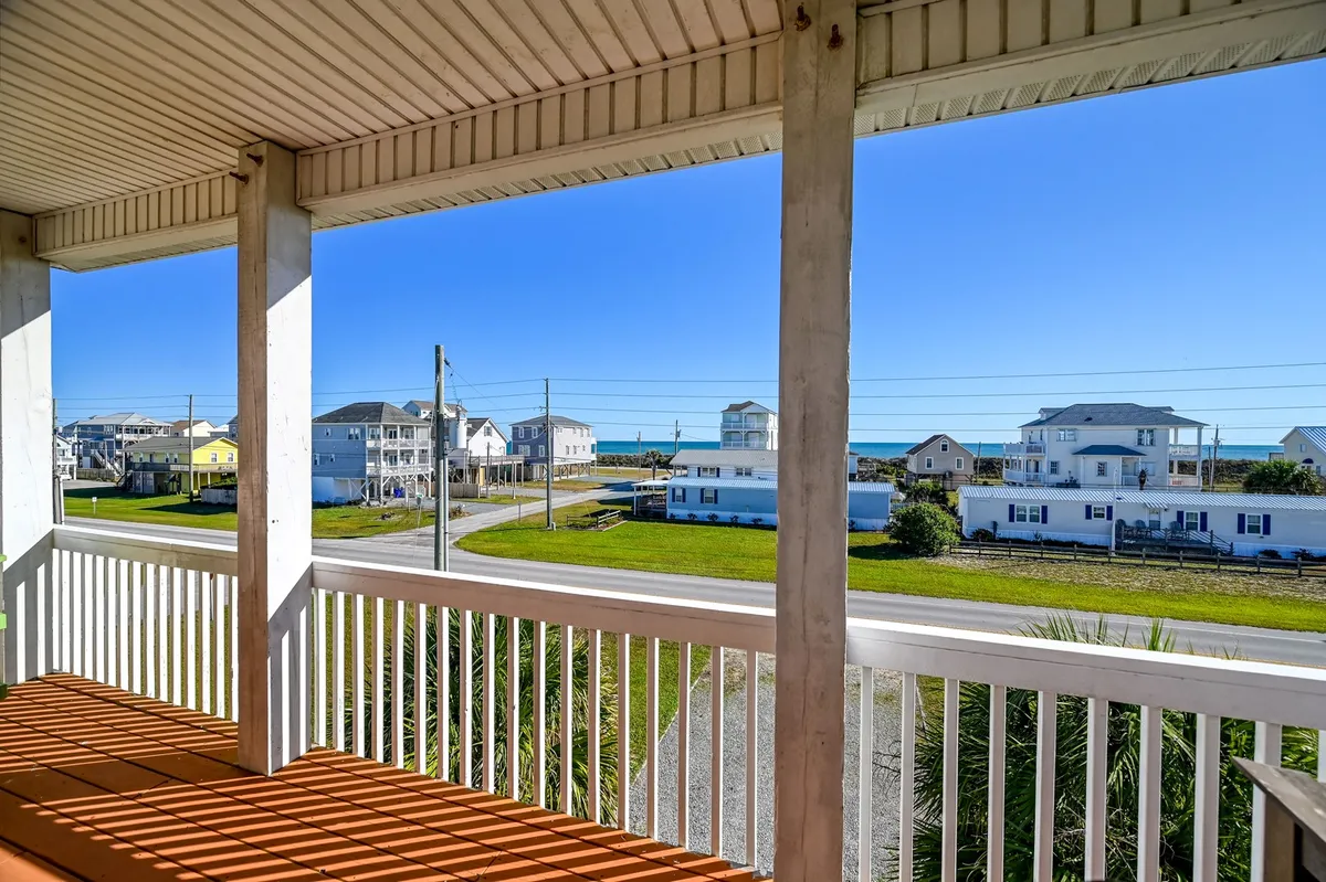 A tranquil porch view with a beach in the background, nearby homes, and a sturdy railing framing the scene. The porch is set up for relaxation and socializing, with comfortable seating for family gatherings, all with a backdrop of sand and sea. The horizon suggests both sunrise coffee moments and evening barbecues.