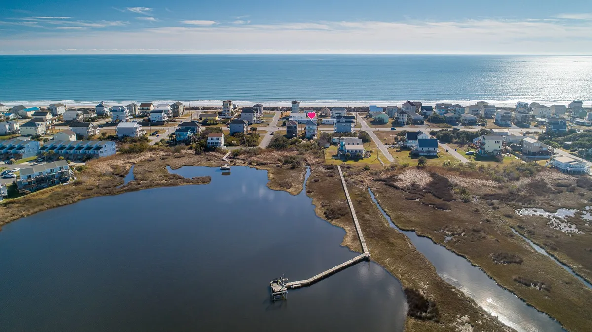 A sweeping view capturing the ocean, houses lining the shore, a prominent pier stretching into the water, and marshland visible nearby. The scene is tranquil, with evidence of human activity like fishing and boating, as well as natural beauty with wildlife habitats in the marsh.